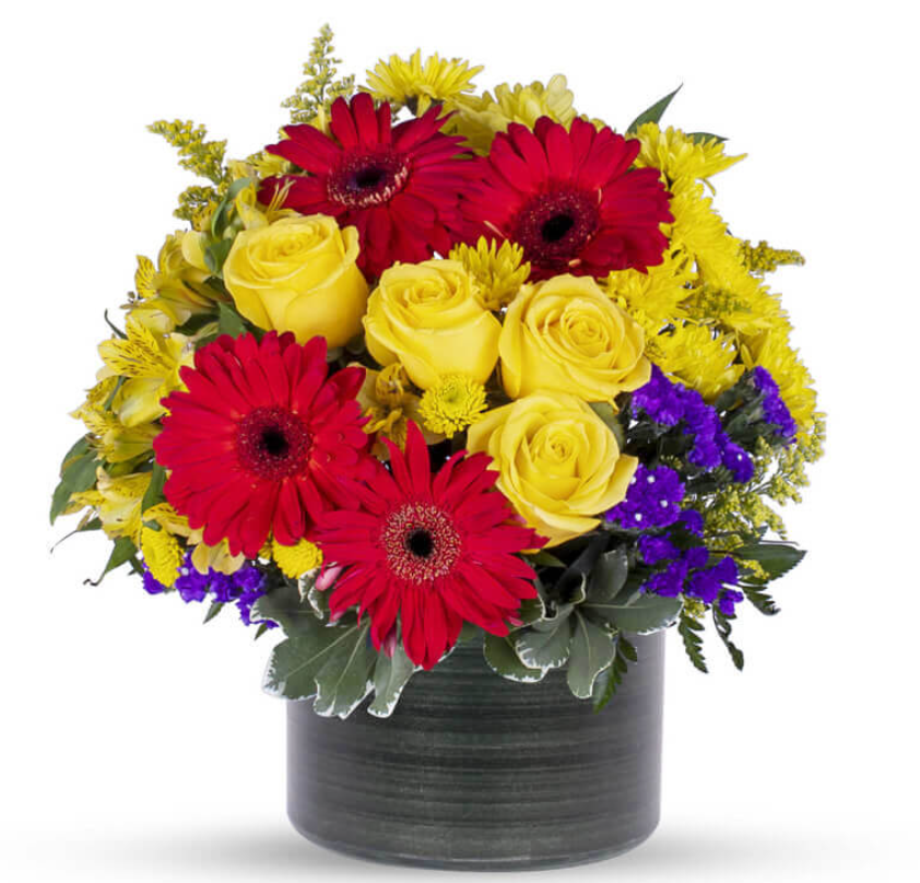 Mixed flower arrangement with red gerbera daisies, yellow roses, purple accents in ceramic vase