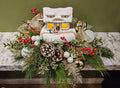 Festive holiday floral arrangement with pinecones, red berries, greenery, and a snow-covered Fudge Shoppe house decoration, designed by Joshua's Greenery Barrie Florist.