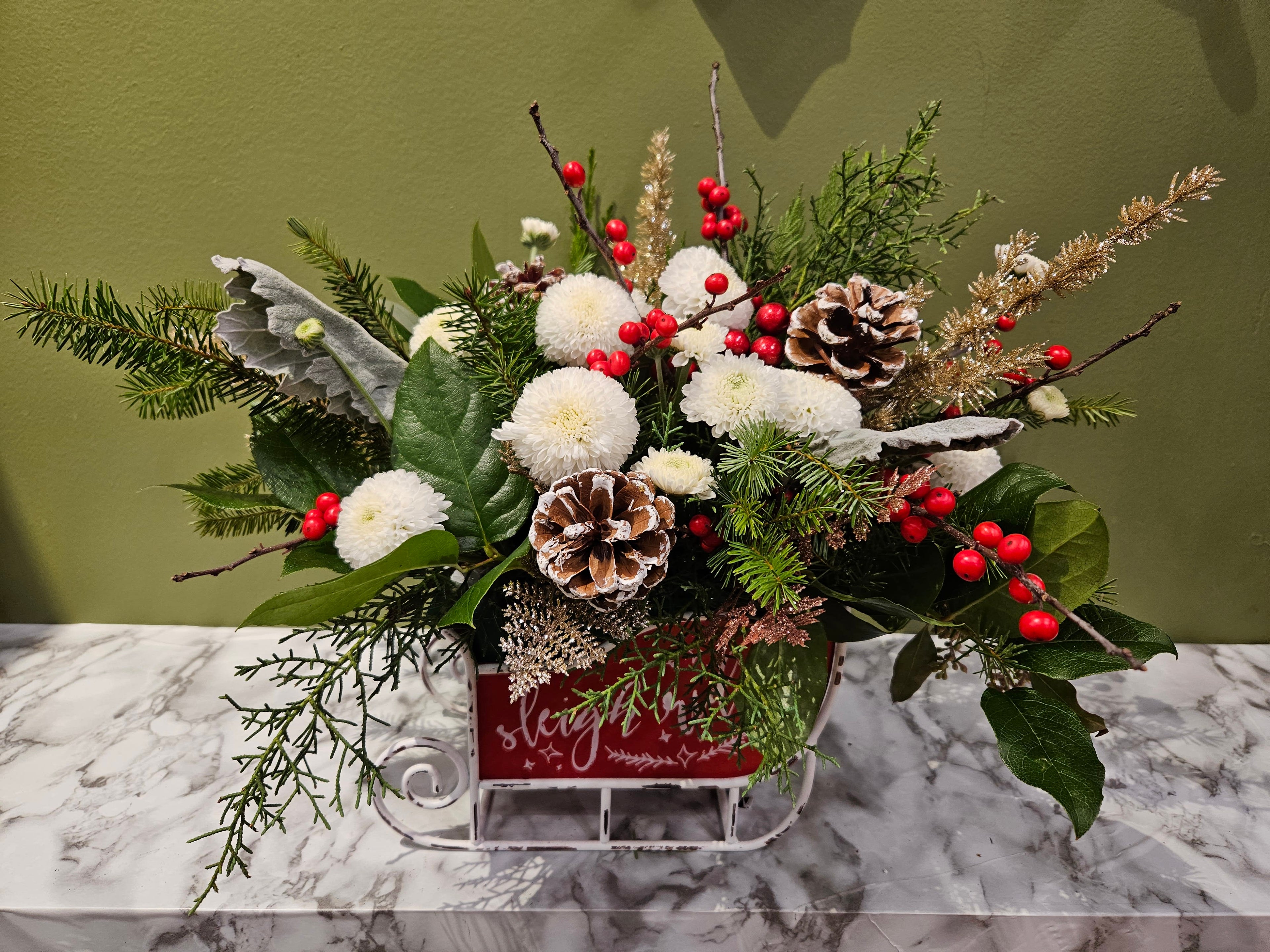 Festive floral arrangement with pinecones, white flowers, red berries, and greenery in a red sleigh, Joshua's Greenery Barrie Florist.
