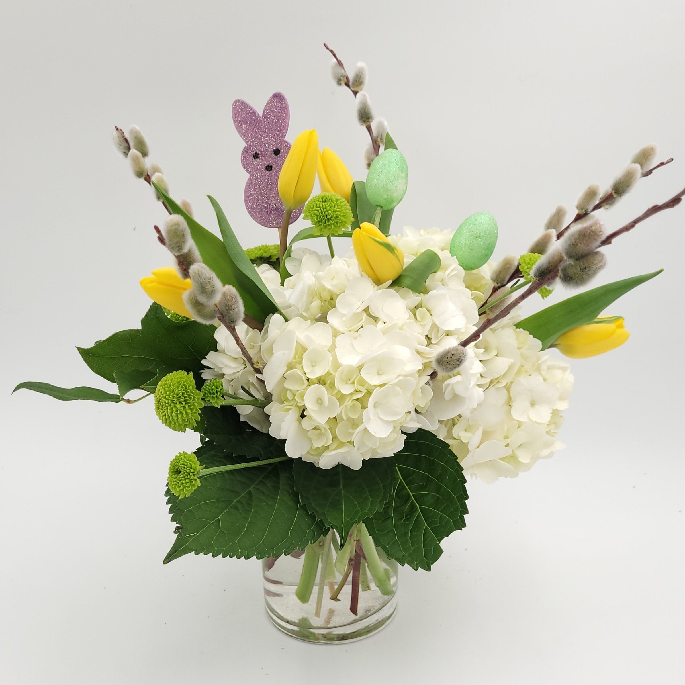 Spring floral arrangement with white hydrangeas, yellow tulips, green accents, and a purple bunny pick in a clear vase from Joshua's Greenery Barrie Florist.